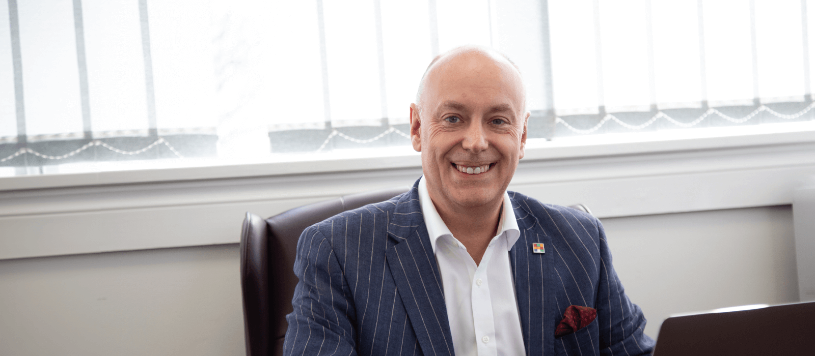 Andrew Scott, dressed in a pinstripe blazer, smiles at his desk at Purplex Marketing, representing his leadership in marketing and business growth.